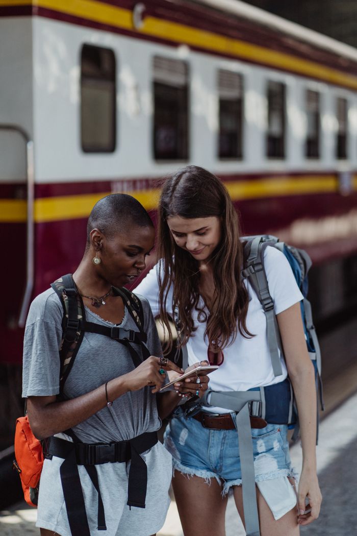 Two women backpackers checking phone at a train station, exploring travel options.